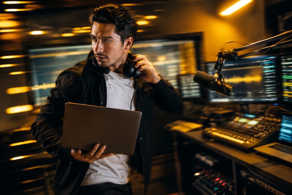 Latino radio broadcaster managing an online radio station with laptop, microphone, and streaming equipment in a fast-paced studio